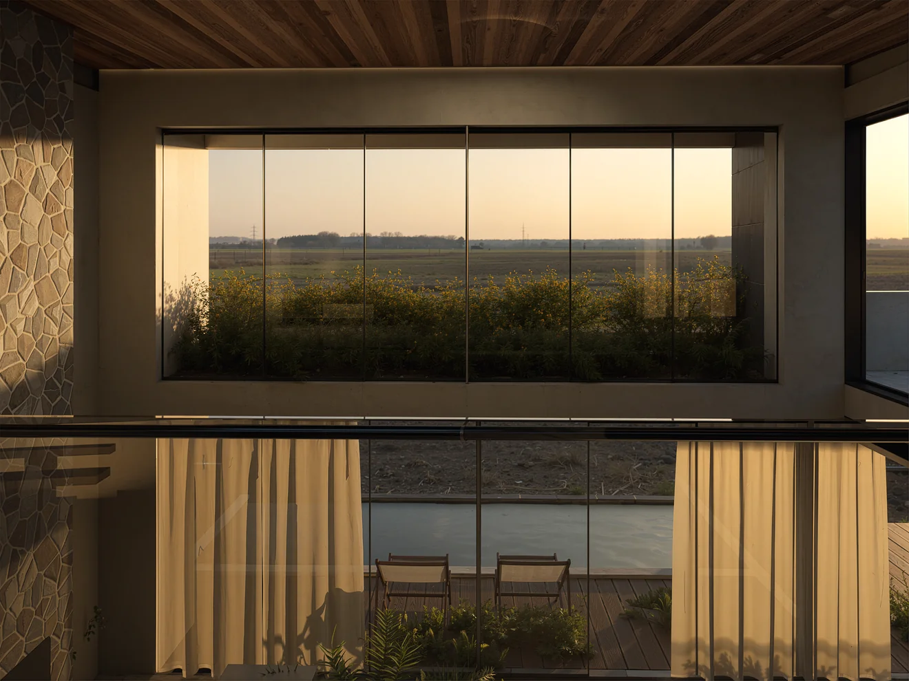 Vista interior de Casa Patio Horizonte con techo de madera, muro revestido en piedra, ventanales y paisaje lejano al atardecer